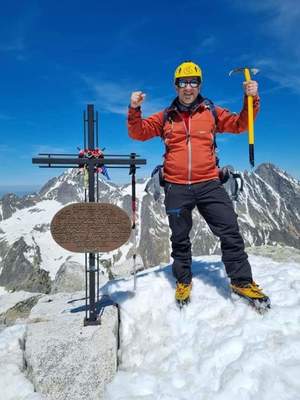 Na zdjęciu widoczny alpinista, który zdobył szczyt, mężczyzna stoi z podniesionymi rękami przy krzyżu.
