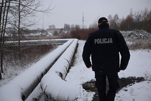 Na zdjęciu umundurowany policjant w scenerii zimowej idzie wzdłuż rur ciepłowniczych.