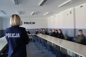 zdjęcie przedstawia policjantkę w mundurze, stojącą tyłem, oraz uczniów klasy mundurowej siedzących  w sali konferencyjnej KMP w Bytomiu